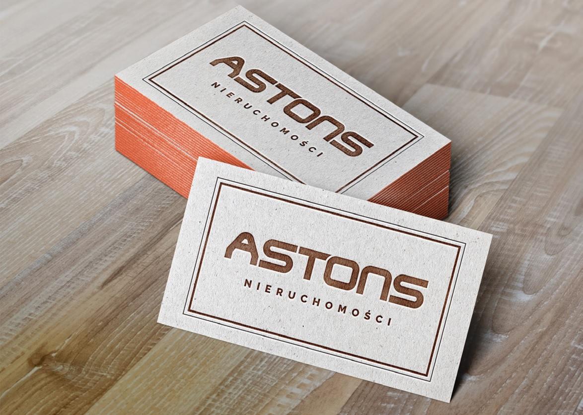 Stos wizytówek firmy Aston's Nieruchomości z pomarańczowymi brzegami, leżących na drewnianym blacie.