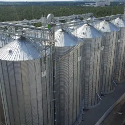 Srebrne, stalowe silosy zbożowe z systemem transportu górnego, widoczne z góry na tle zielonego lasu i zabudowań przemysłowych.