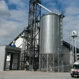 Kompleks silosów zbożowych ze stalową konstrukcją wsporczą, widoczny biały van na tle błękitnego nieba z chmurami.