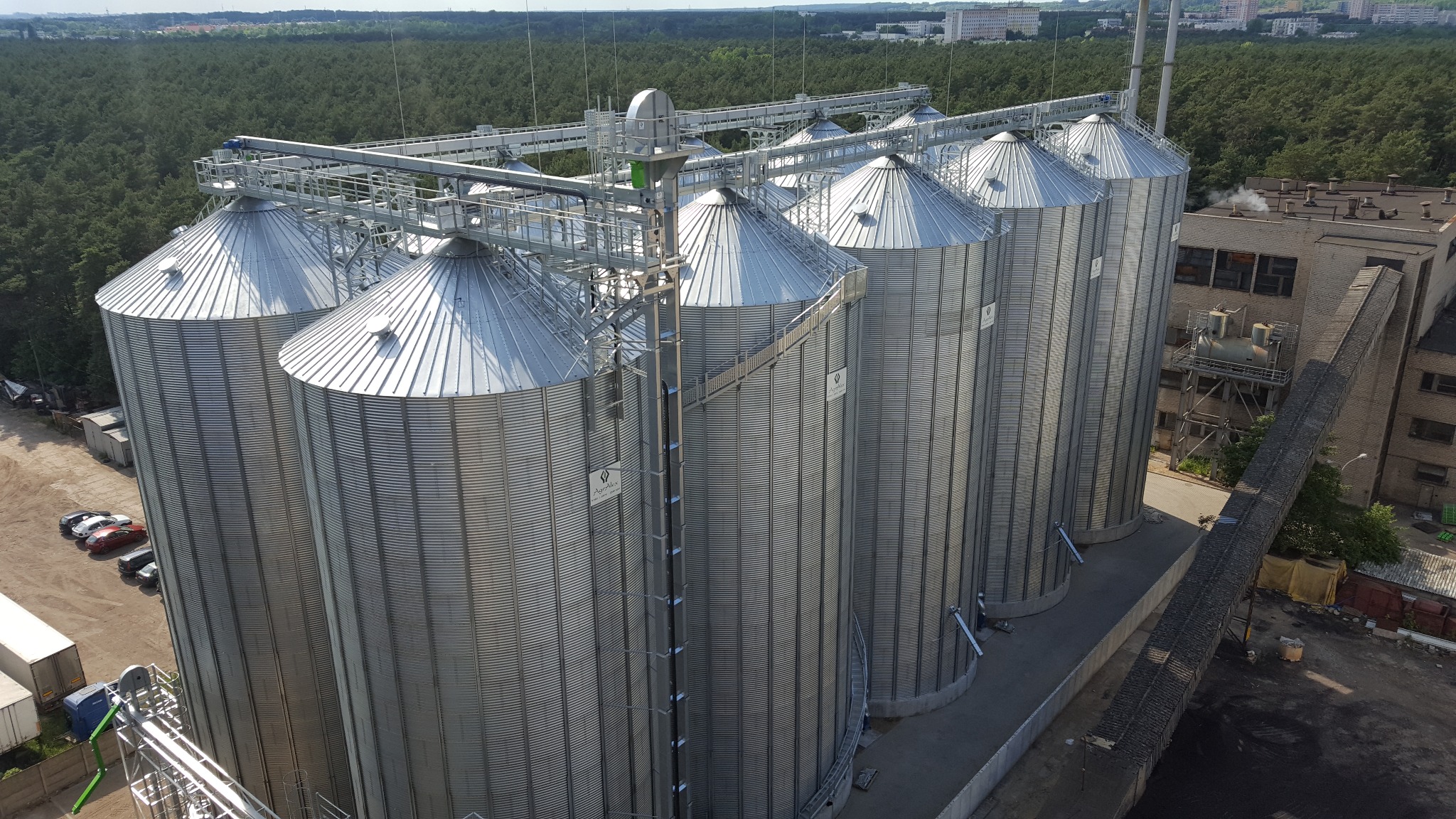 Srebrne, stalowe silosy zbożowe z systemem transportu górnego, widoczne z góry na tle zielonego lasu i zabudowań przemysłowych.