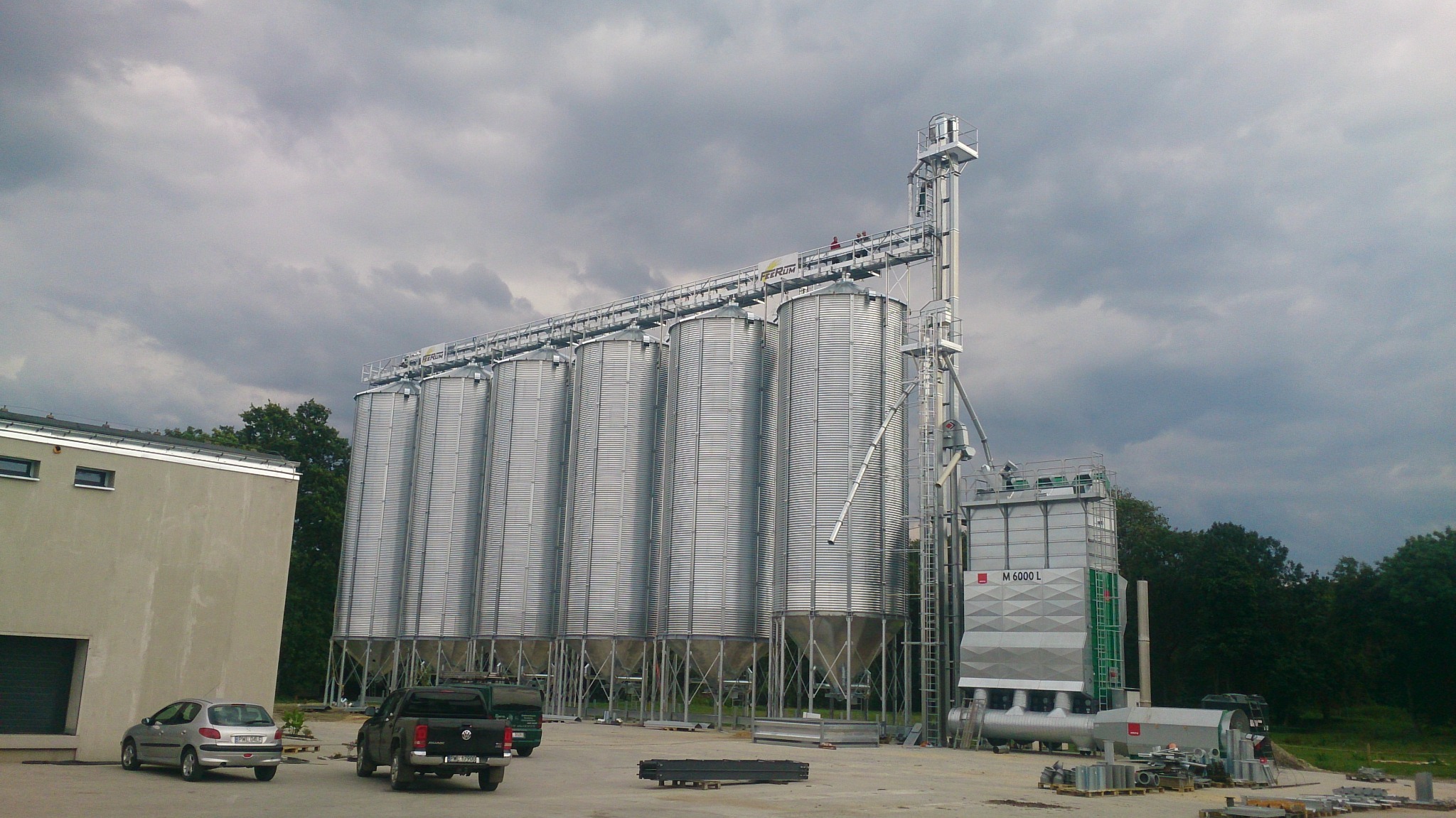 Widok na zespół silosów zbożowych z podajnikiem, zlokalizowanych na placu z zaparkowanymi samochodami osobowymi i dostawczymi, na tle pochmurnego nieba.
