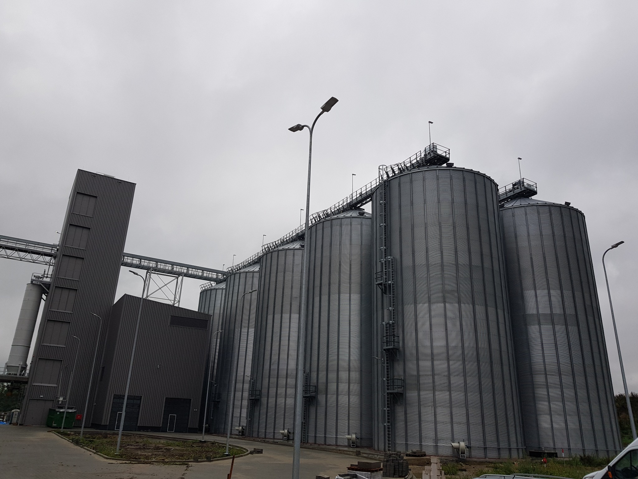 Kompleks silosów zbożowych o metalowej, falistej konstrukcji, połączonych pomostami, z budynkiem obsługi i oświetleniem ulicznym na tle pochmurnego nieba.