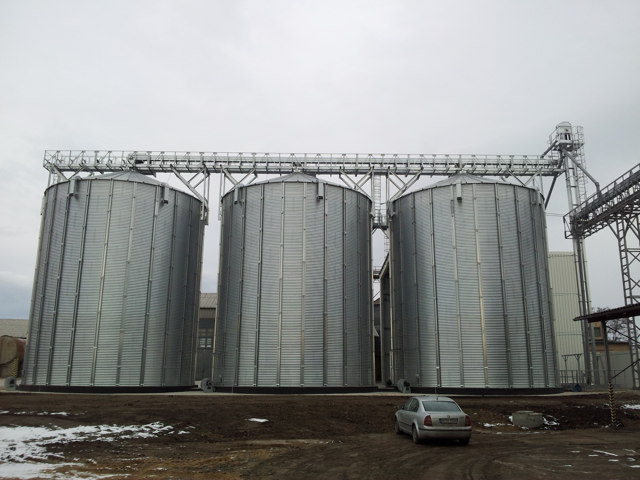 Trzy metalowe silosy zbożowe połączone konstrukcją stalową, z widocznym fragmentem instalacji transportowej, na tle pochmurnego nieba i ziemi z resztkami śniegu, obok zaparkowany srebrny samochód.