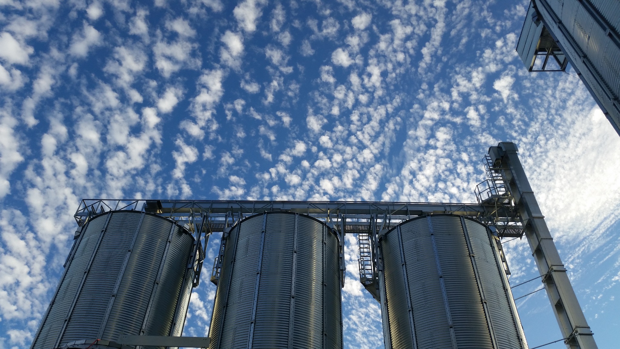Trzy wysokie, metalowe silosy z pomostami i drabinkami, sfotografowane z dołu na tle błękitnego nieba z białymi, kłębiastymi chmurami.