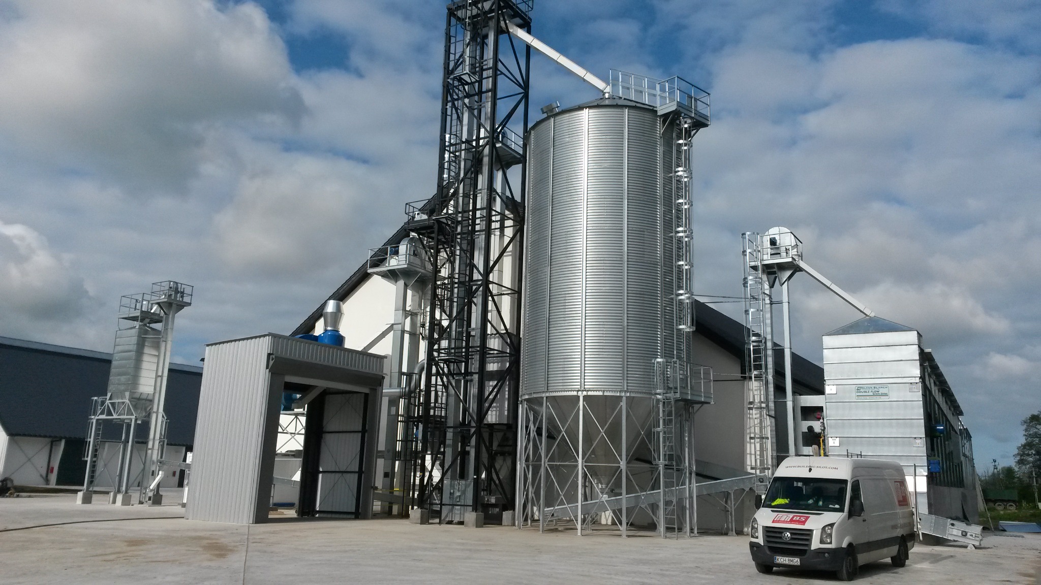 Kompleks silosów zbożowych ze stalową konstrukcją wsporczą, widoczny biały van na tle błękitnego nieba z chmurami.