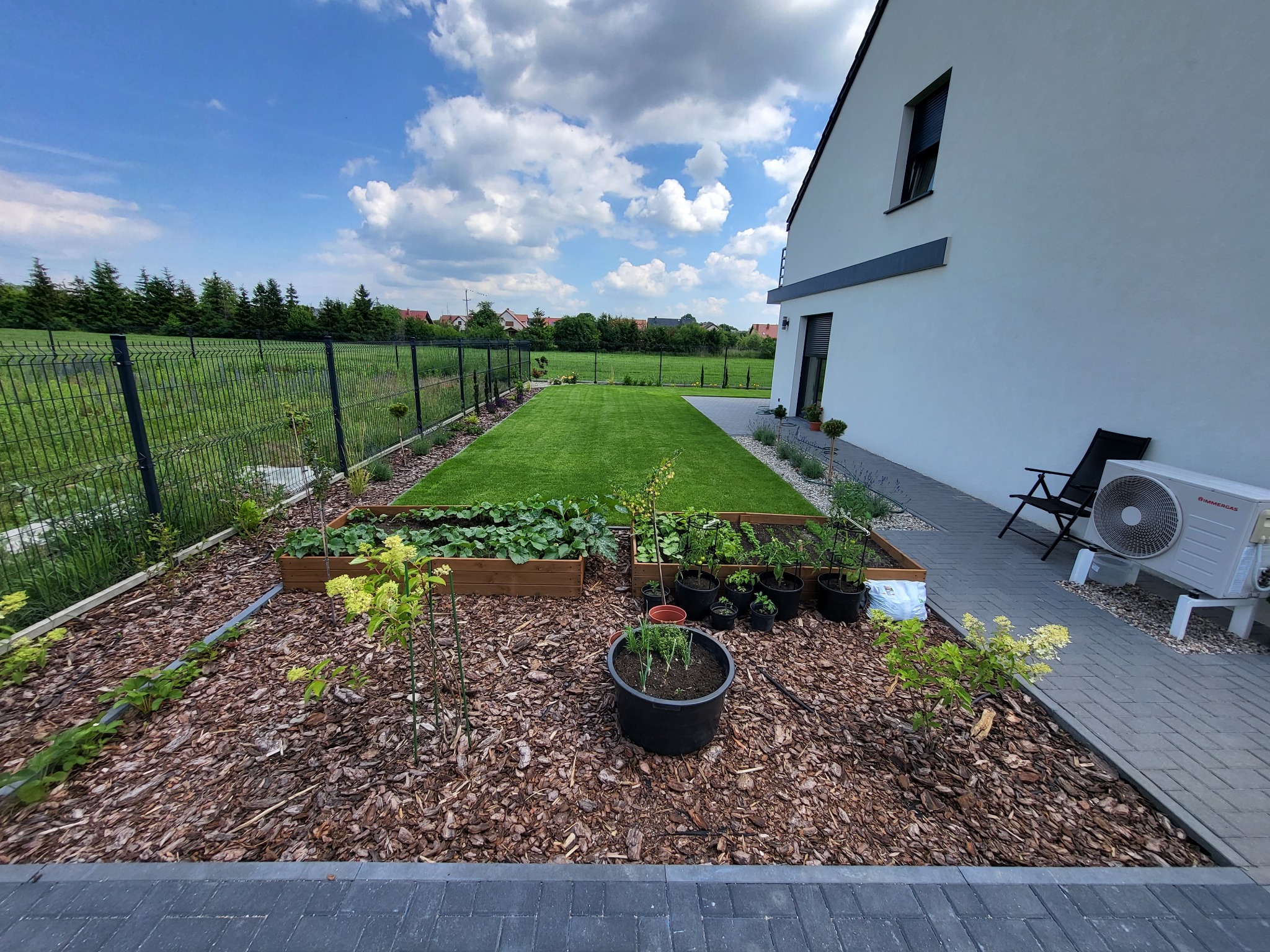 Uporządkowany ogród przydomowy z rabatami warzywnymi w drewnianych skrzyniach, doniczkami z ziołami i równo przystrzyżonym trawnikiem, wyściółkowany korą, na tle nowoczesnego domu z klimatyzatorem.