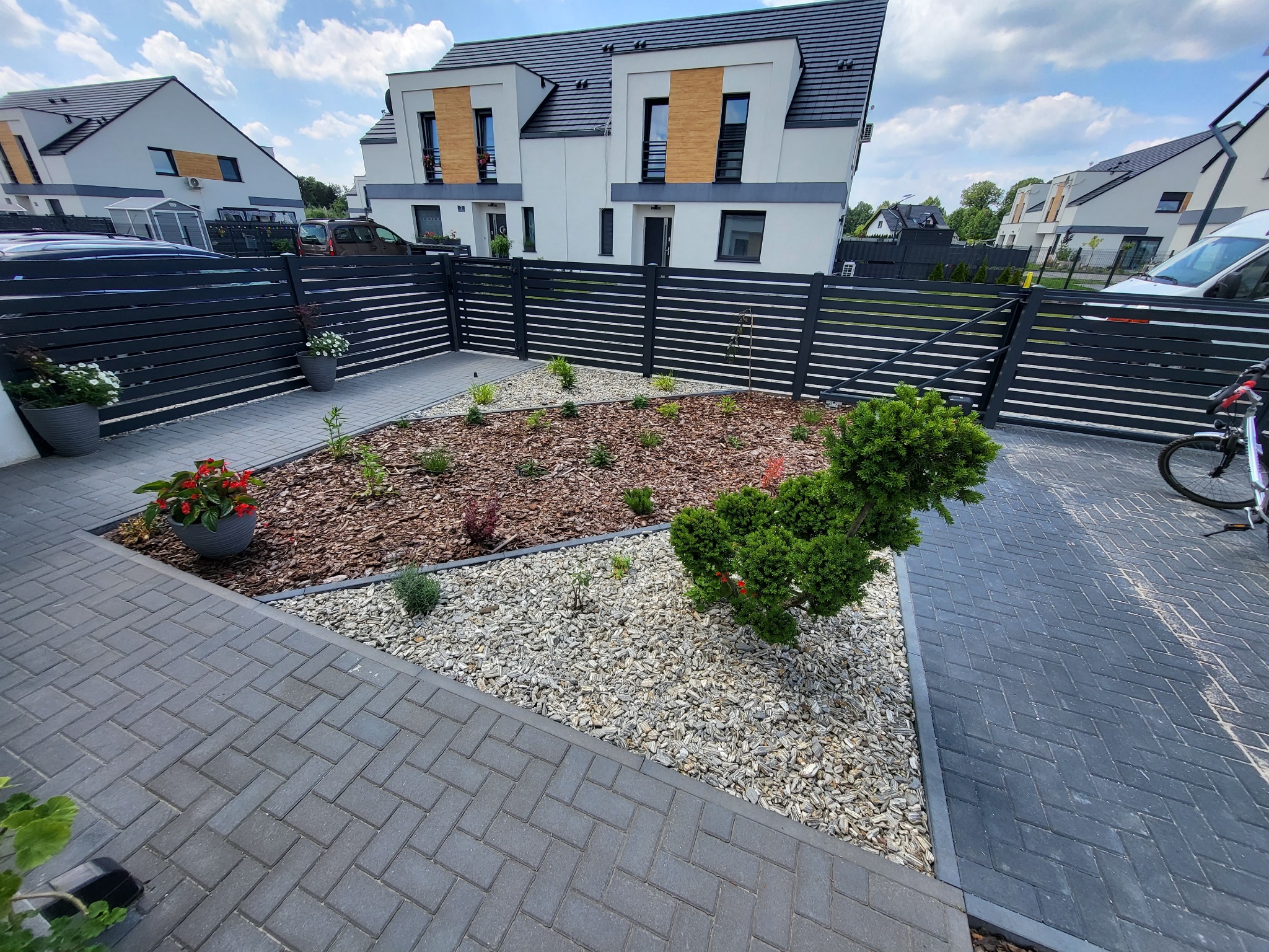 Nowoczesny ogród przed domem z geometrycznymi rabatami wysypanymi korą i białym żwirem, niskie krzewy ozdobne, trawnik w budowie, czarne ogrodzenie panelowe, kostka brukowa w odcieniach szarości.