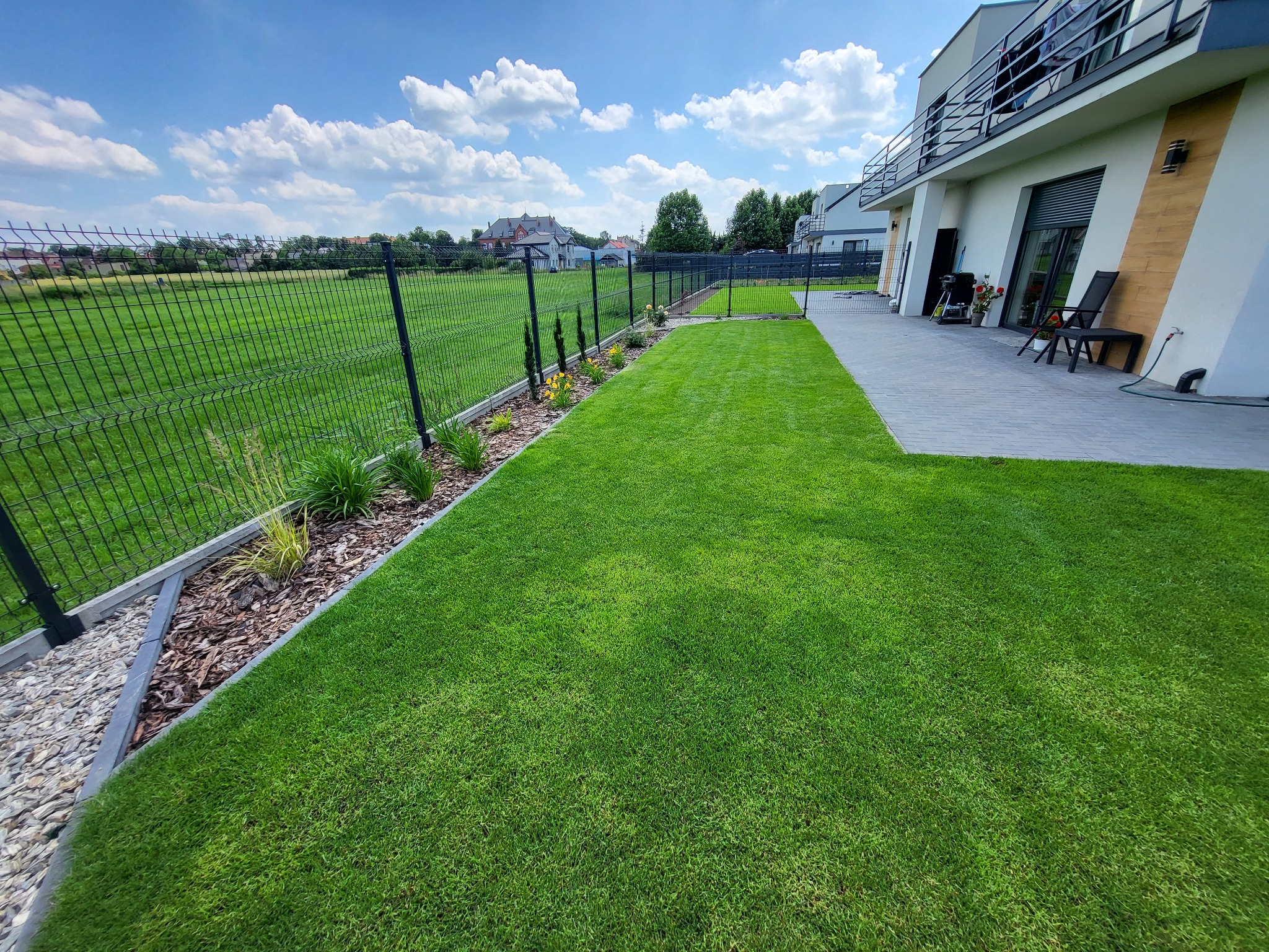 Zadbany trawnik z rolki w Wąsoszu, widok na nowoczesny dom z tarasem i czarnym ogrodzeniem panelowym, rabaty z korą i roślinami ozdobnymi.