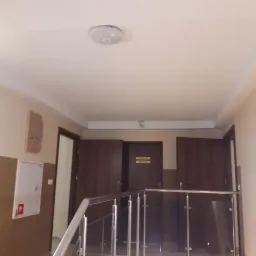 Korytarz biurowy z pomalowanymi ścianami w odcieniach beżu, z widocznymi drzwiami do biur, oświetleniem sufitowym i metalową barierką schodową.