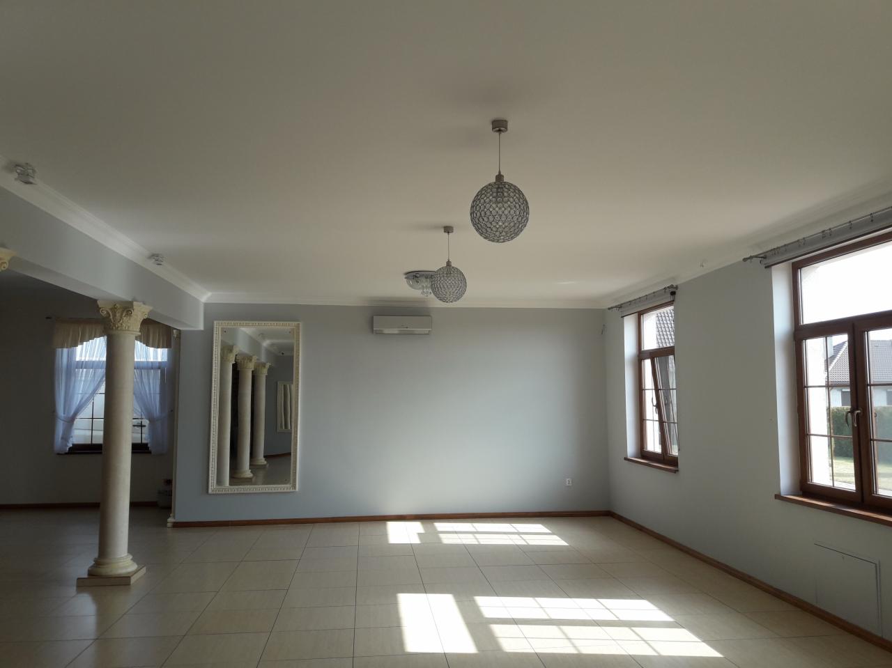 Duże, puste pomieszczenie z szarą ścianą, kolumnami dekoracyjnymi, lustrem z kolumnami, trzema kulistymi lampami sufitowymi i brązowymi ramami okiennymi, przygotowane pod aranżację.