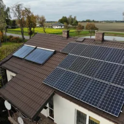 Dom jednorodzinny z brązowym dachem, na którym zamontowano panele słoneczne i kolektory słoneczne, widok z góry na tle krajobrazu wiejskiego.