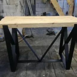 Stół z blatem z jasnego drewna i czarnymi, metalowymi nogami o geometrycznym kształcie w warsztacie.