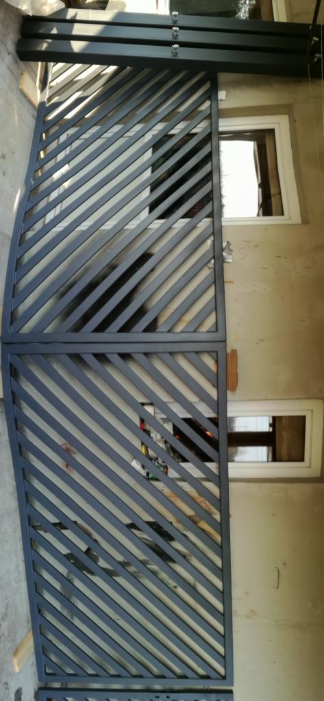 Dwa segmenty metalowego ogrodzenia z ukośnymi prętami, przygotowane do montażu, opierają się o ścianę budynku z widocznymi dwoma oknami.