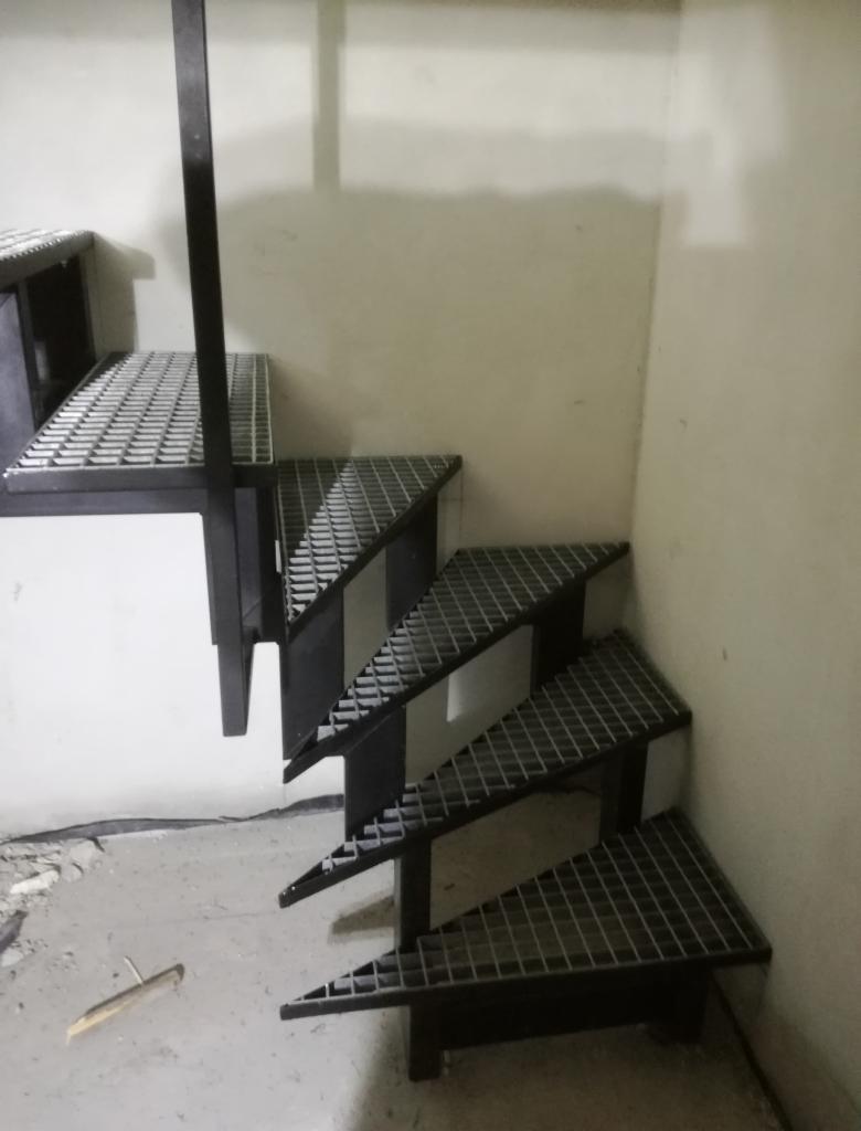 Metalowe schody kratowe z czarnymi wspornikami zamontowane w rogu pomieszczenia o jasnych ścianach, widoczne ślady po budowie na podłodze.