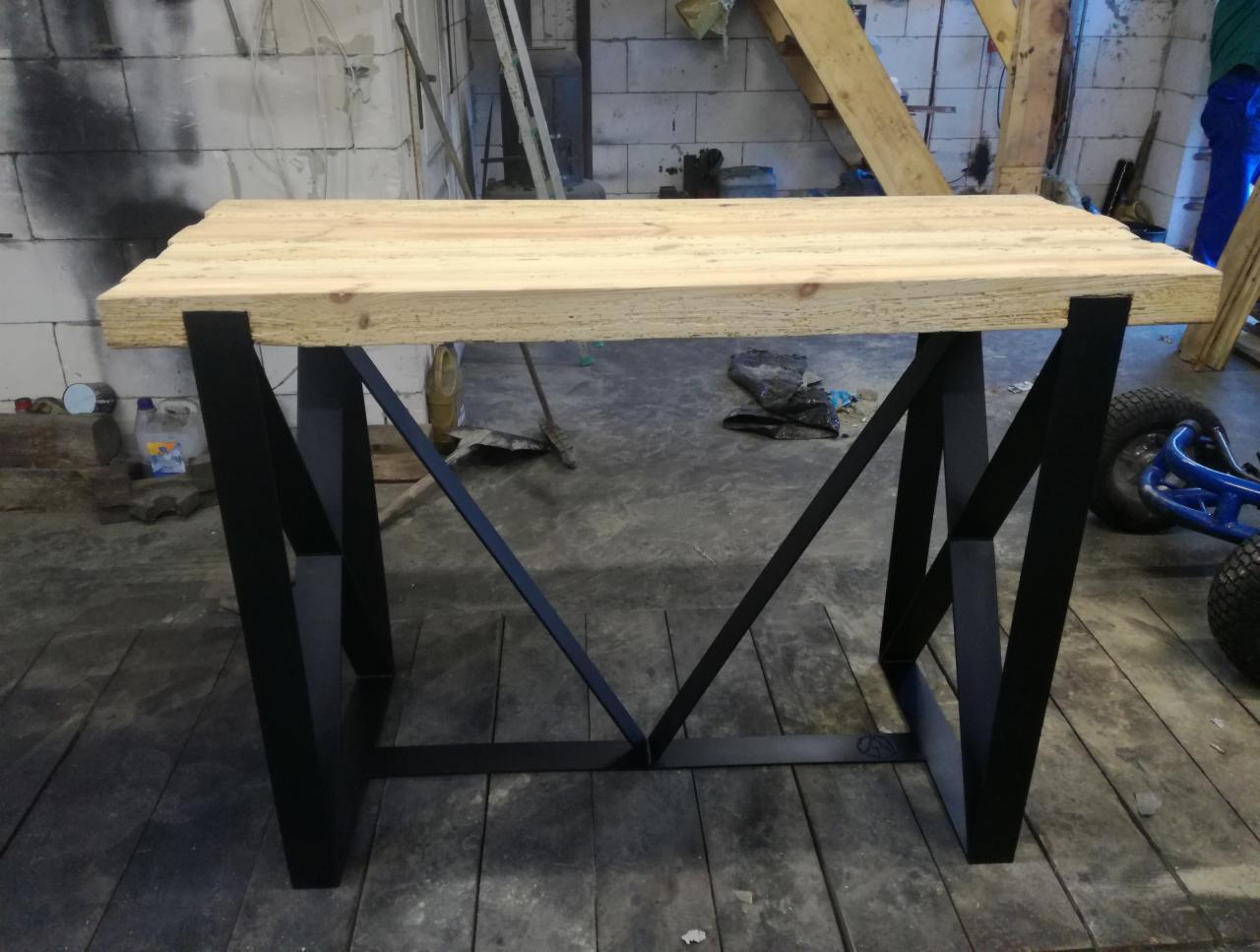 Stół z blatem z jasnego drewna i czarnymi, metalowymi nogami o geometrycznym kształcie w warsztacie.