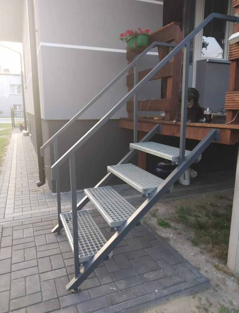 Zewnętrzne schody metalowe z kratowymi stopniami prowadzące na drewniany taras z kwiatami i psem.