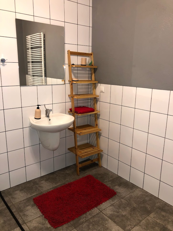 Łazienka z umywalką na postumencie, drewnianą drabinką-regałem, czerwonym dywanikiem i białymi kafelkami z szarą ścianą w tle.