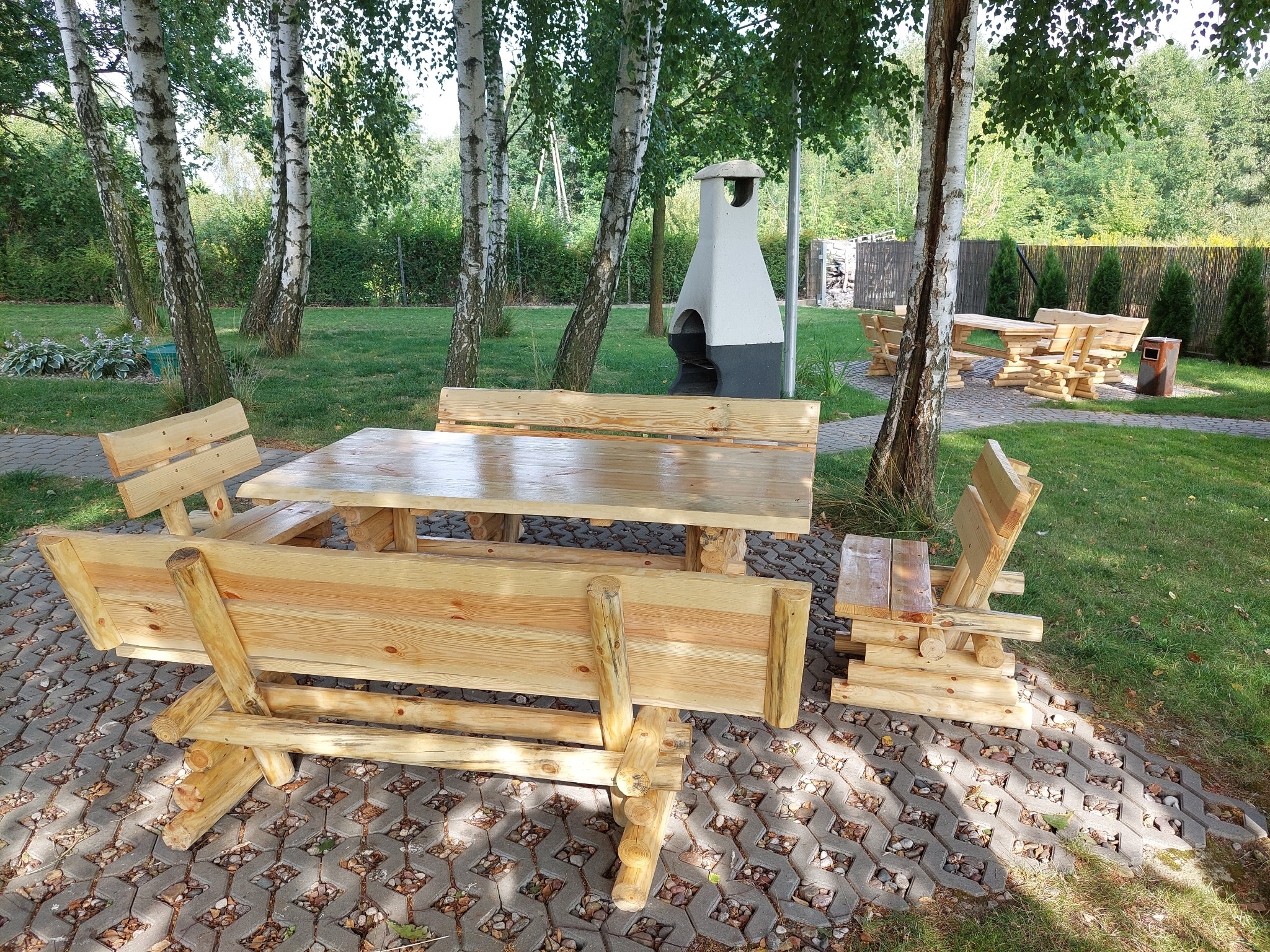 Drewniany zestaw mebli ogrodowych z bali: stół i ławy z oparciami, na tle trawnika, drzew i grilla murowanego.