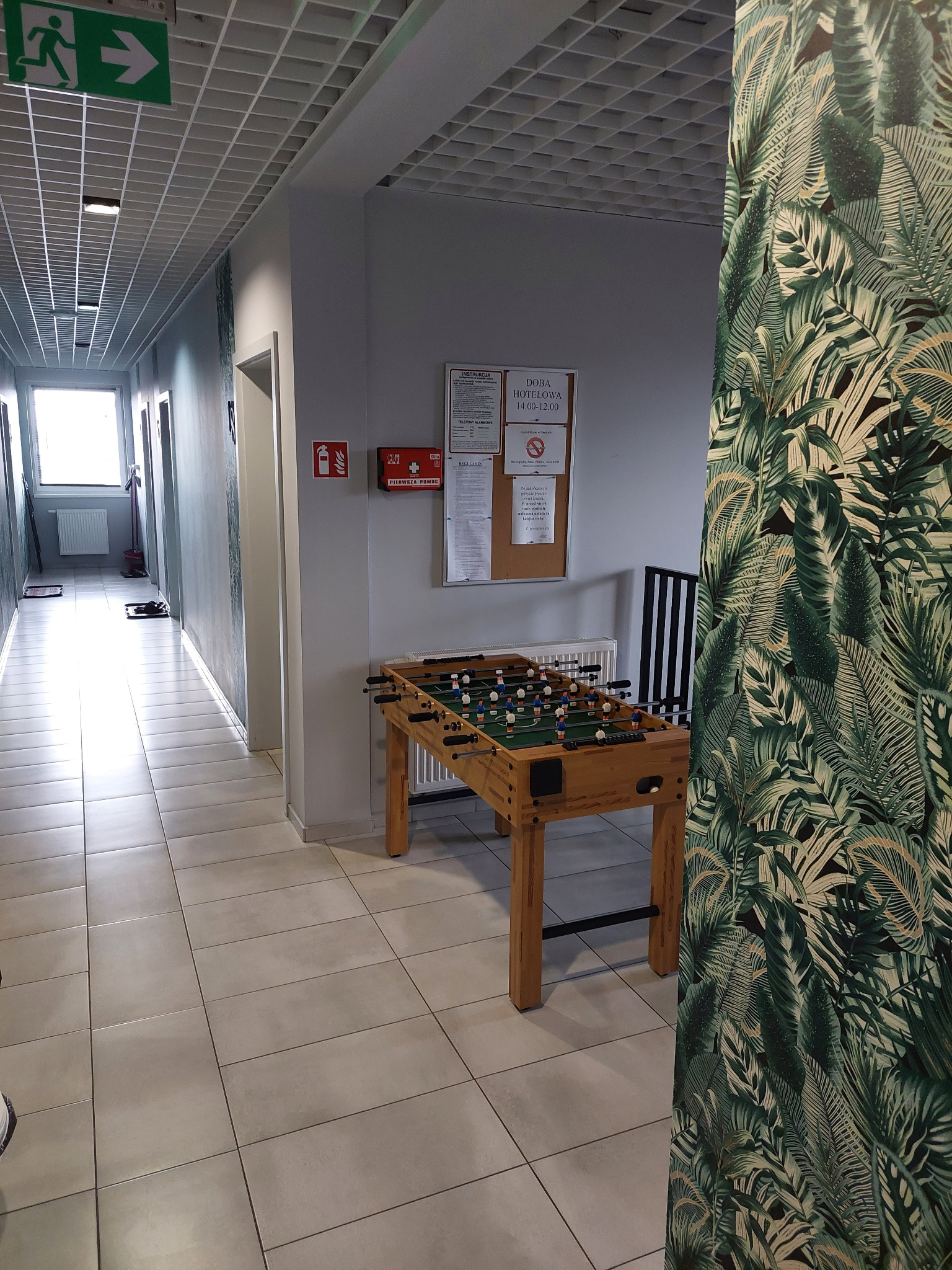 Korytarz z grą w piłkarzyki, apteczką pierwszej pomocy, plakatem informacyjnym i tapetą w tropikalne liście w tle.