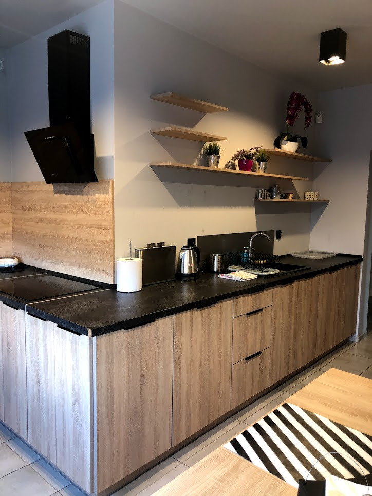 Jasna kuchnia z drewnianymi szafkami, czarnym blatem i półkami z roślinami doniczkowymi.