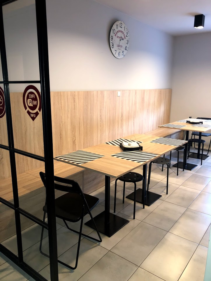 Wnętrze lokalu gastronomicznego z długimi, drewnianymi stołami z czarno-białymi podkładkami, czarnymi krzesłami i zegarem na ścianie, widoczne przez szklaną ścianę z czarnymi ramami.