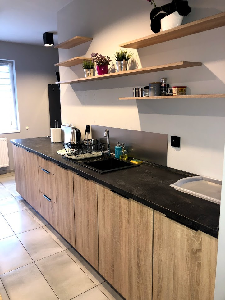 Nowoczesna kuchnia z drewnianymi szafkami, ciemnym blatem i otwartymi półkami z roślinami i akcesoriami kuchennymi.