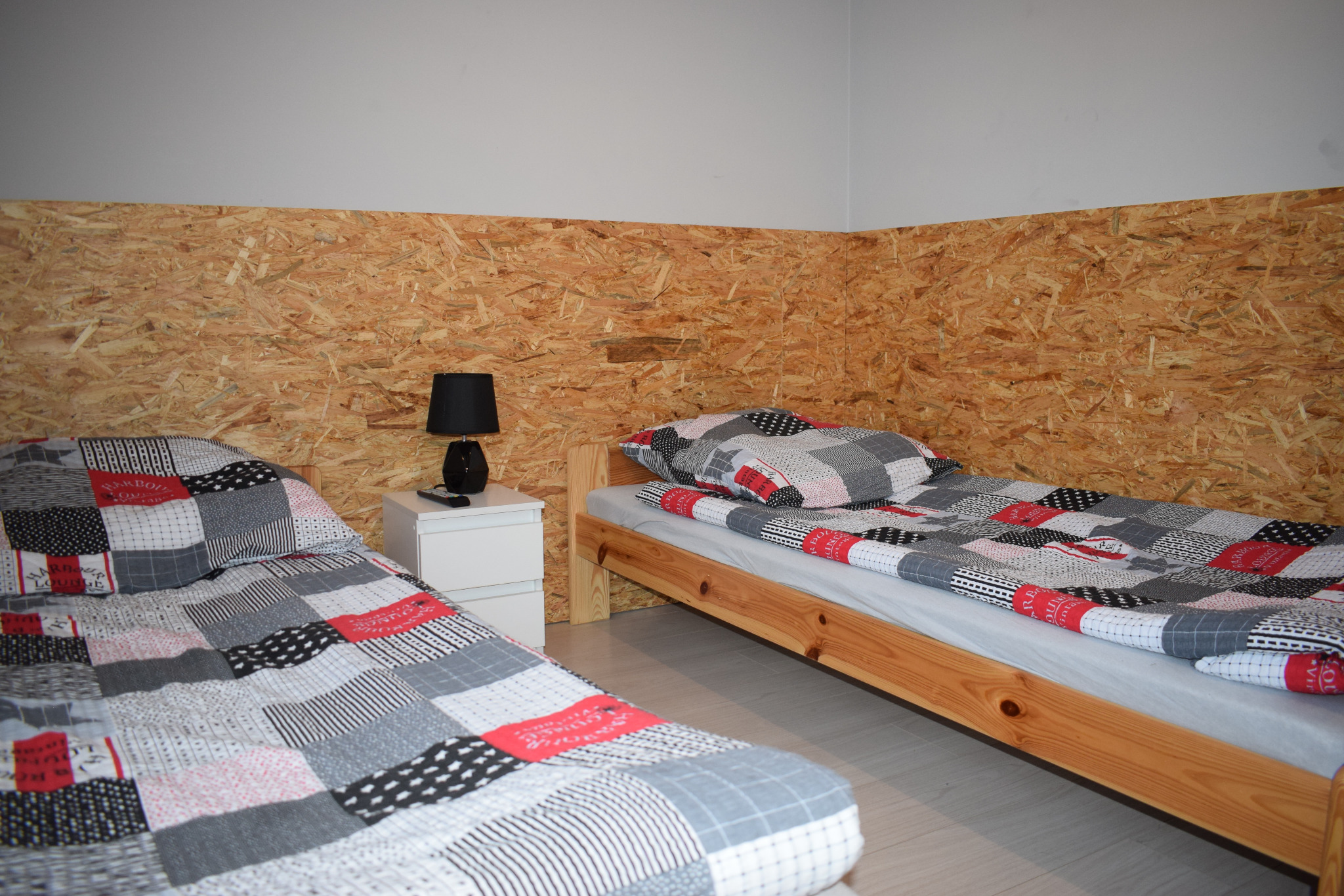 Dwa łóżka pojedyncze z patchworkową pościelą w pokoju z ścianą wykończoną płytami OSB, mała czarna lampka nocna na białej szafce.
