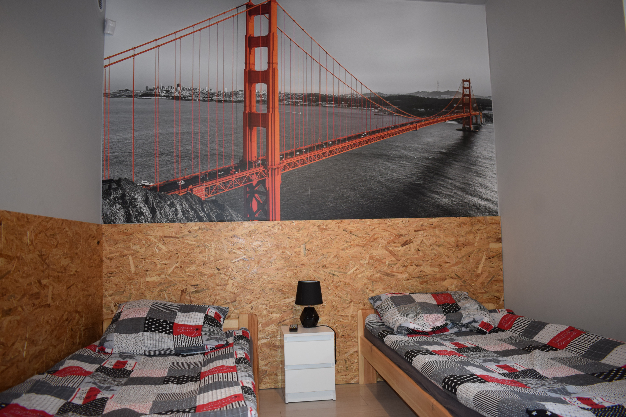 Pokój z dwoma łóżkami pojedynczymi, drewnianą ścianą z płyty OSB i fototapetą przedstawiającą most Golden Gate.