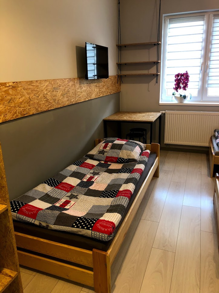 Pokój z pojedynczym łóżkiem w drewnianej ramie, patchworkową kołdrą, biurkiem z czarnymi nogami, wiszącymi półkami i telewizorem na ścianie wyłożonej płytą OSB.