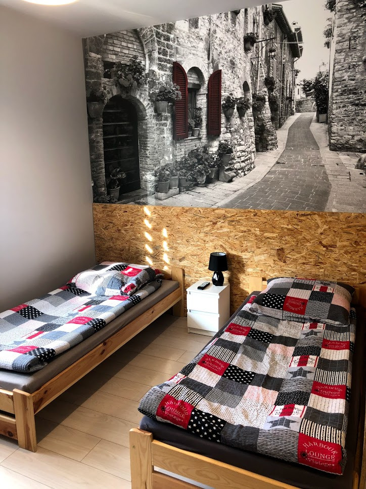 Pokój z dwoma łóżkami pojedynczymi, nad którymi umieszczono fototapetę przedstawiającą uliczkę w stylu włoskim z kamiennymi budynkami i czerwonymi okiennicami, z widocznym fragmentem drewnianego...