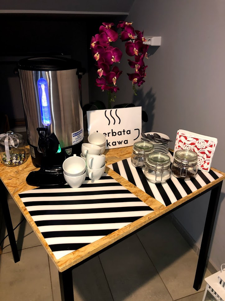 Stół z czarno-białym bieżnikiem, na którym stoi automat do grzania wody, stos filiżanek, słoiczki z zawartością i serwetnik, a w tle purpurowe orchidee.