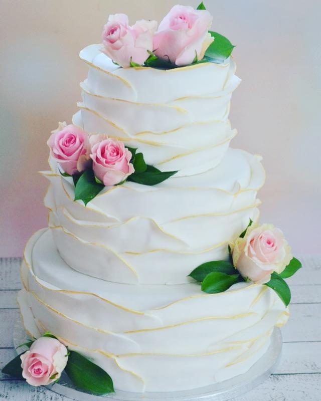Trzypiętrowy tort weselny z białym lukrem, złoconymi akcentami i dekoracją z różowych róż oraz zielonych liści na drewnianym stole.
