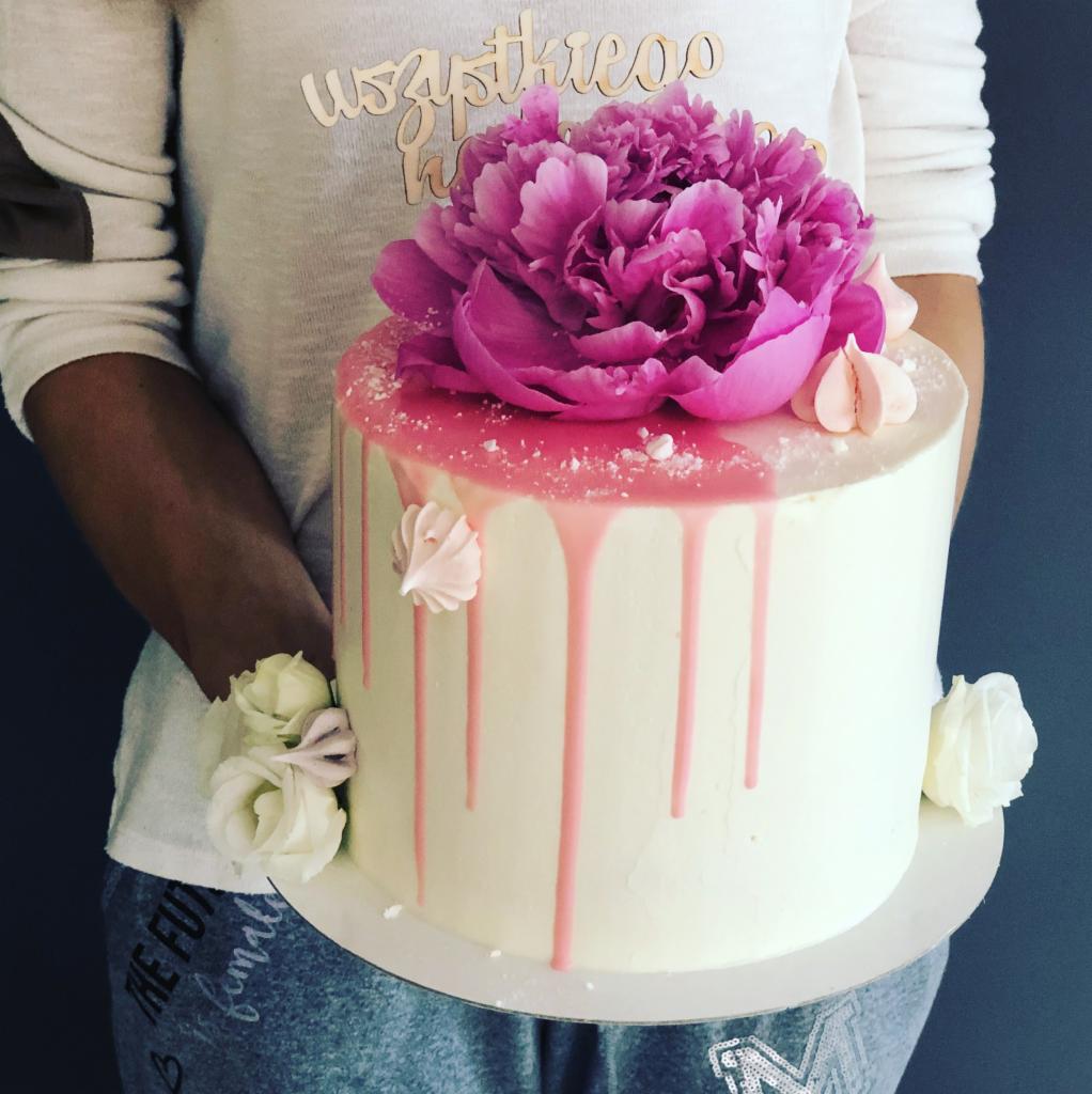 Okrągły tort z białym kremem, różowym lukrem spływającym po bokach, ozdobiony dużym, różowym kwiatem piwonii i białymi bezami, trzymany przez osobę w szarej bluzie i dresach.