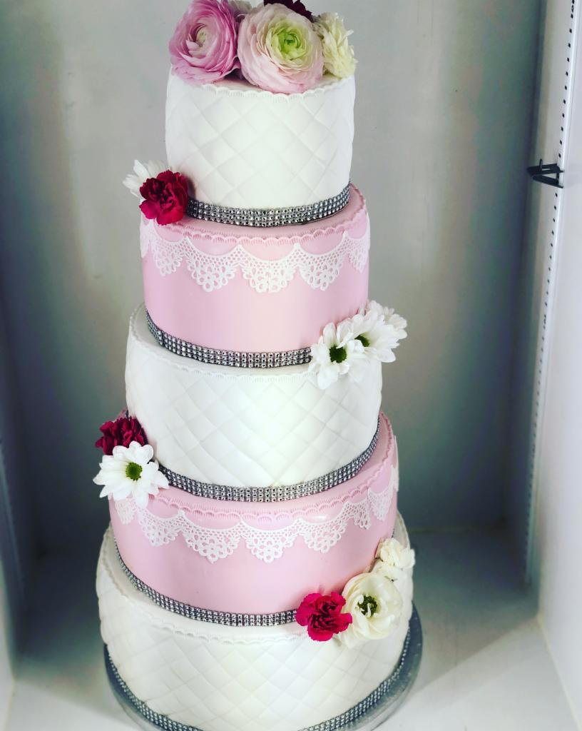 Piętrowy tort weselny w odcieniach bieli i różu, zdobiony kwiatami, imitacją pikowania i srebrnymi pasami.