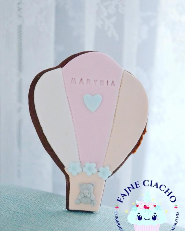 Piernik w kształcie balonu z imieniem Marysia i misiem, dekorowany lukrem w pastelowych kolorach, na tle jasnej tkaniny.