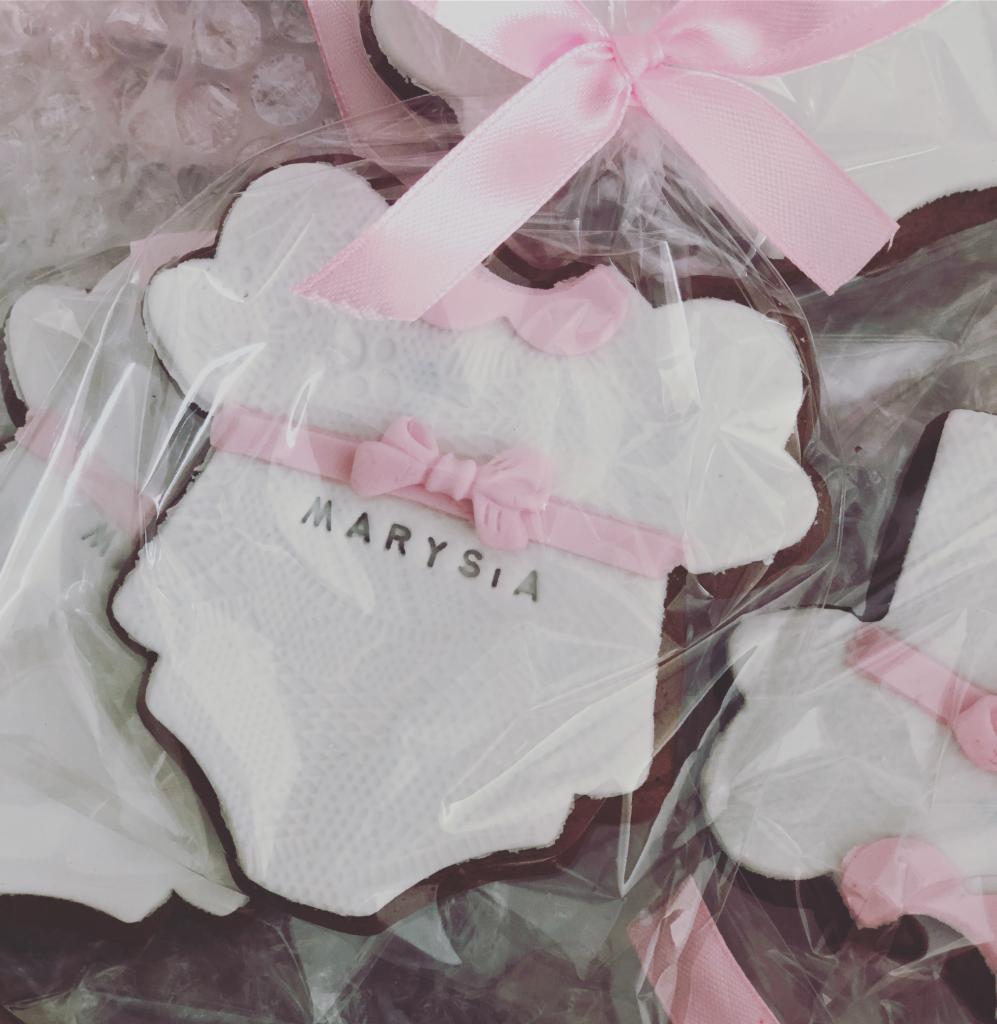 Pierniczki w kształcie niemowlęcych bodów z białym lukrem, różową wstążką i napisem 'Marysia', zapakowane w folię z różową kokardką.