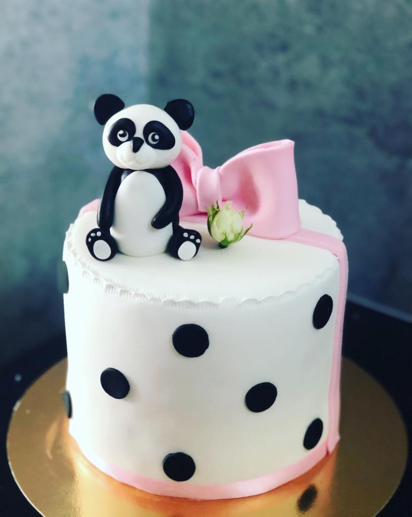 Tort urodzinowy z białym lukrem, czarnymi kropkami, różową wstążką, różową kokardą i figurką pandy na górze, stojący na złotym podkładzie.