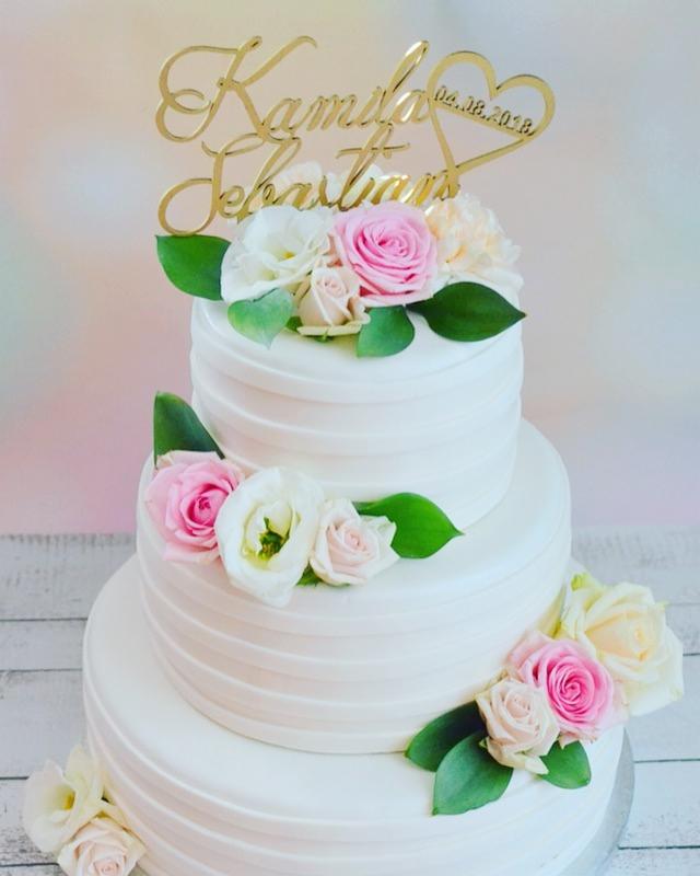 Trzypiętrowy tort weselny z białym kremem, dekorowany różowymi i białymi różami oraz złotym topperem z imionami 'Kamila i Sebastian' i datą 04.08.2018.