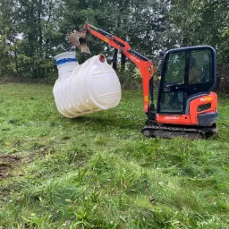 Pomarańczowa mini koparka Kubota podnosi biały, plastikowy zbiornik z niebieskimi elementami na tle zielonej trawy i drzew