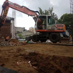 Pomarańczowy koparko-ładowarka Liebherr 900 pracuje na placu budowy, w tle mur z cegły i konstrukcja zbrojeniowa, operator w kabinie w kamizelce odblaskowej, pracownik przy gruzowisku.