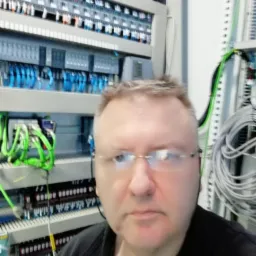 Mężczyzna w okularach na tle szafy sterowniczej z przewodami i modułami, prezentujący elementy instalacji elektrycznej w rozdzielnicy.