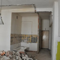 NAAM-BUD - Wnętrze w trakcie rozbiórki: odsłonięte cegły, resztki tynku, gruz. Widoczna wanna i wisząca lampa. Ściany częściowo pomalowane na żółto.