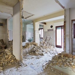 NAAM-BUD - Wnętrze mieszkania w trakcie rozbiórki: gruz na podłodze, usunięte fragmenty ścian, widoczne instalacje. Drabina oparta o ścianę. Widok na łazienkę z sanitariatami.