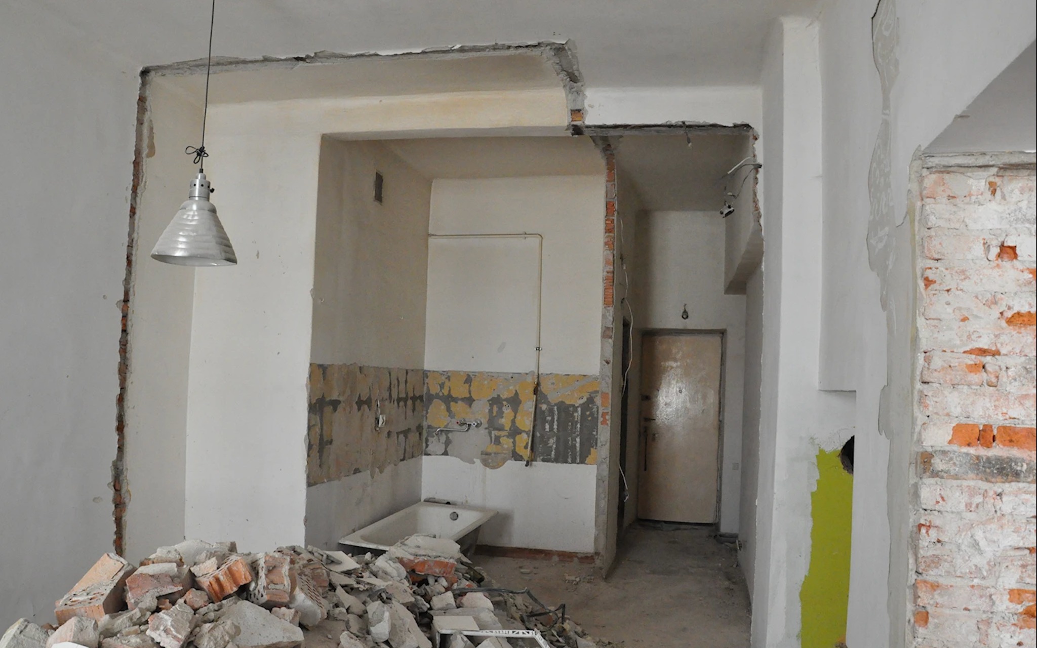 Wnętrze w trakcie rozbiórki: odsłonięte cegły, resztki tynku, gruz. Widoczna wanna i wisząca lampa. Ściany częściowo pomalowane na żółto.