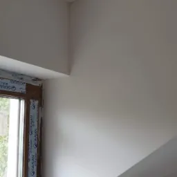 Wnętrze po malowaniu, widoczne świeżo pomalowane ściany i sufit w jasnym odcieniu, z oknem w lewym dolnym rogu, którego rama jest jeszcze zabezpieczona folią.