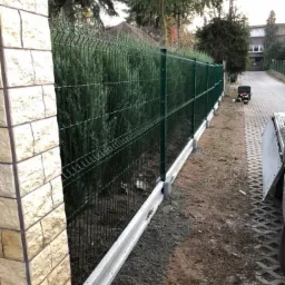 Zielone ogrodzenie panelowe z podmurówką betonową, widok z boku, tło z tujami i budynkiem, narzędzia na chodniku w oddali.