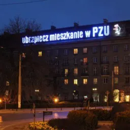 Wielopiętrowy budynek oświetlony w nocy z neonowym napisem 'Ubezpiecz mieszkanie w PZU' na dachu.