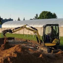 Żółta mini koparka Yanmar SV18 w trakcie prac ziemnych, wykop pod fundamenty, widoczny wykopany rów i pryzmy ziemi, w tle szklarnia.