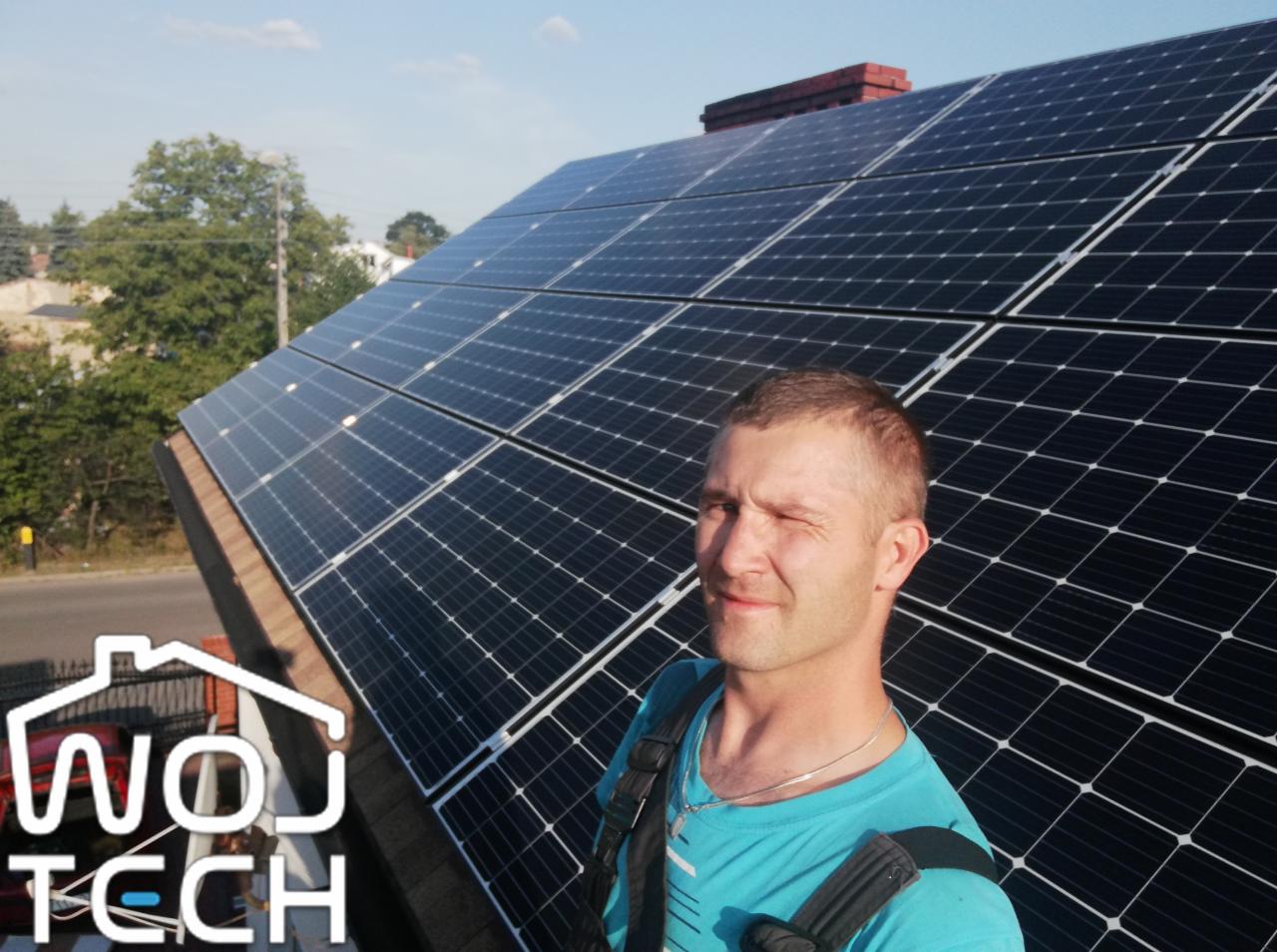 Uśmiechnięty instalator z przymrużonym okiem na tle paneli fotowoltaicznych na dachu. Widoczne logo firmy WOJTECH. Słoneczny dzień, instalacja ukończona.