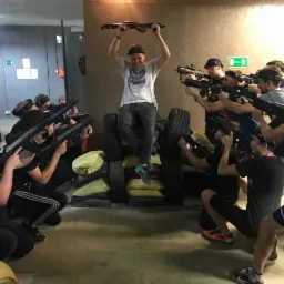 Grupa osób celuje z pistoletów laserowych w pomieszczeniu z oponami i poduszkami; jedna osoba siedzi na stosie opon i trzyma pistolet nad głową.